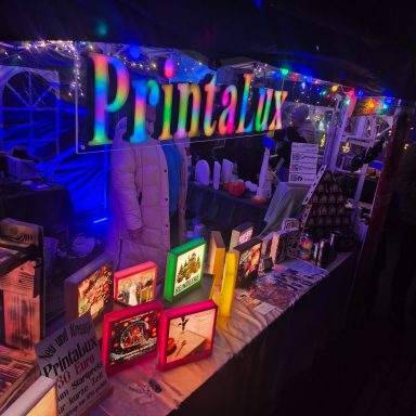 Bunte Lichter und ausgestellte Bilder auf einem Marktstand von PrintaLux.