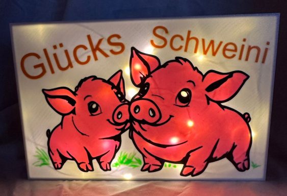 Zwei rosa Ferkel mit leuchtender Schriftzug "Glücks Schweini" auf einem Schild.